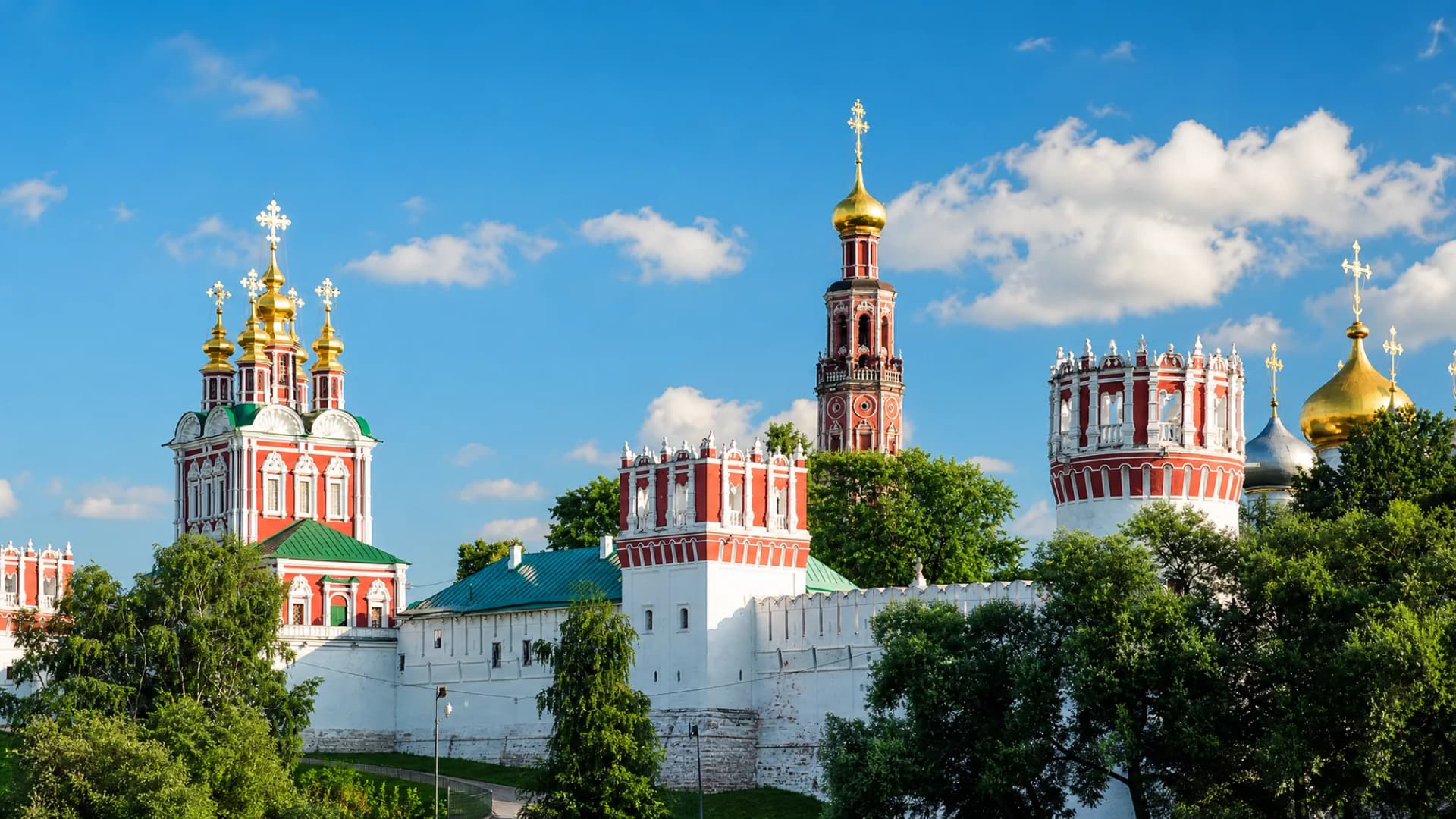 Новодевичий монастырь летним днём: белые стены, красная колокольня и золотые купола над густой зеленью